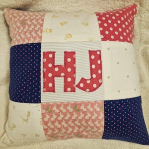 May include: A square patchwork cushion featuring a grid of nine fabric squares with various patterns including pink bunnies, white polka dots, navy blue dots, and stars. The centre square displays the appliqued red polka dot letters HJ.