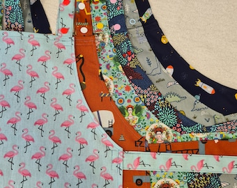 May include: A stack of five colourful aprons with different patterns. The top apron is light blue with pink flamingos, the second is orange with a construction theme, the third is green with a floral pattern, the fourth is grey with a dinosaur pattern, and the bottom apron is navy blue with a rocket ship pattern.