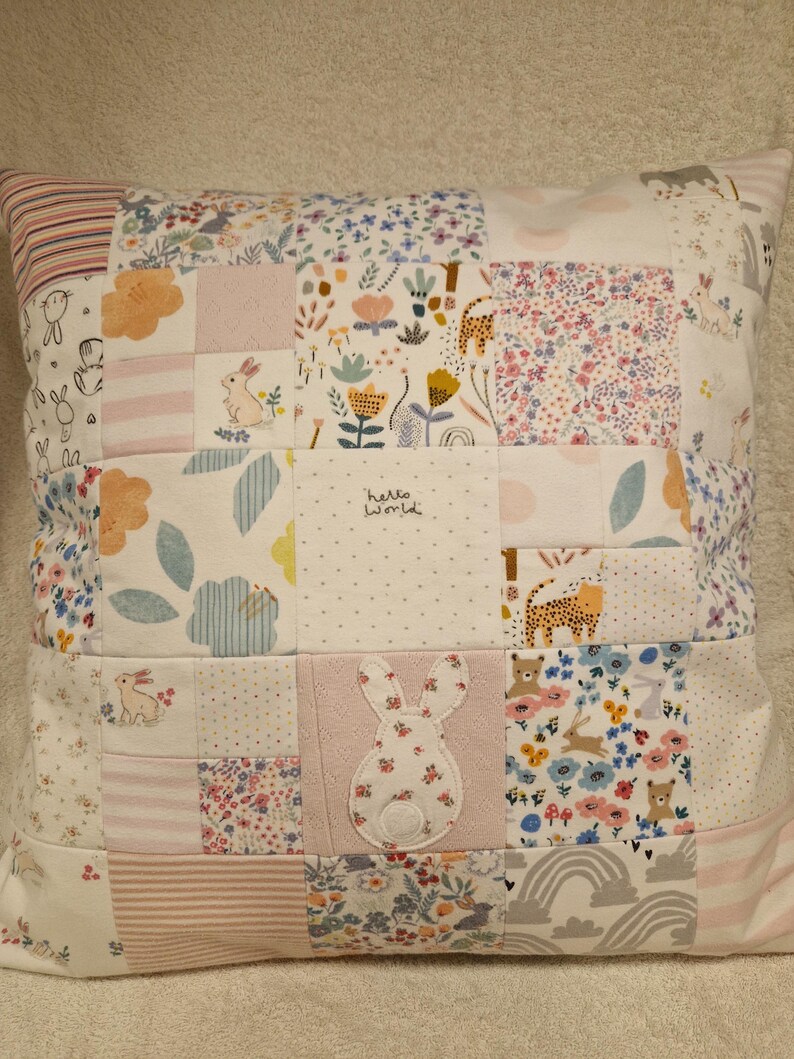 May include: A square patchwork cushion featuring a variety of fabric squares with floral, animal, and striped patterns in soft pastel colours. One square displays the text hello world, and a central square features a white rabbit appliqu&eacute;.