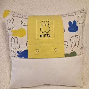 May include: A rectangular throw cushion featuring a white fabric base with a repeating Miffy rabbit pattern in black outlines, yellow, blue, and green. A central yellow fabric band displays a square patch with the Miffy logo and text.