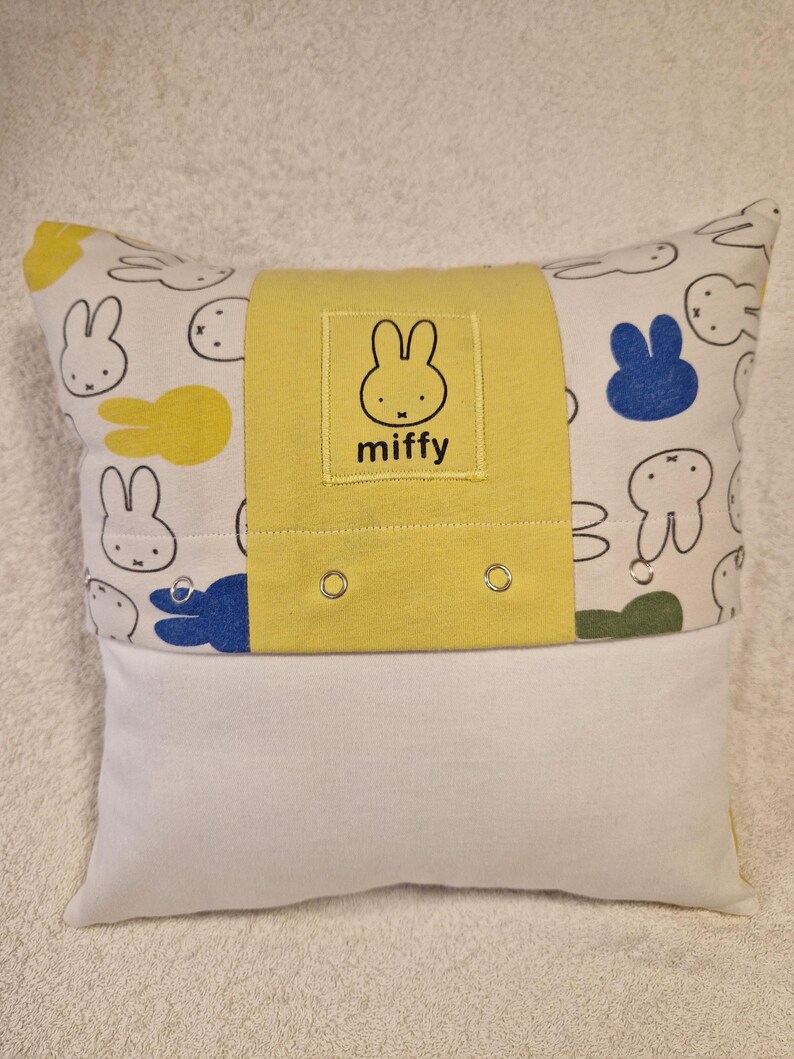 May include: A rectangular throw cushion featuring a white fabric base with a repeating Miffy rabbit pattern in black outlines, yellow, blue, and green. A central yellow fabric band displays a square patch with the Miffy logo and text.