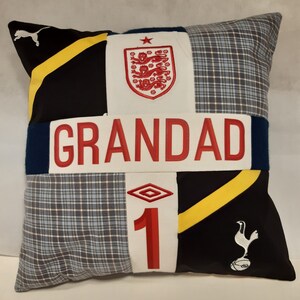 May include: A square patchwork cushion featuring a central white horizontal band with the red text GRANDAD. The design includes black fabric with yellow stripes, blue plaid panels, the England national team crest, and the Tottenham Hotspur logo.