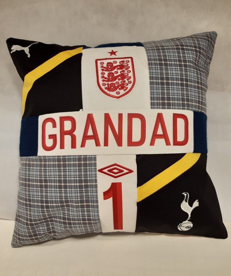 May include: A square patchwork cushion featuring a central white horizontal band with the red text GRANDAD. The design includes black fabric with yellow stripes, blue plaid panels, the England national team crest, and the Tottenham Hotspur logo.