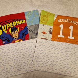 May include: Two rectangular fabric pillowcases featuring patchwork designs. The left case displays Superman with yellow text, while the right case features an orange panel with the text NEDERLAND 11. Both have white sections with small cloud and moon patterns.