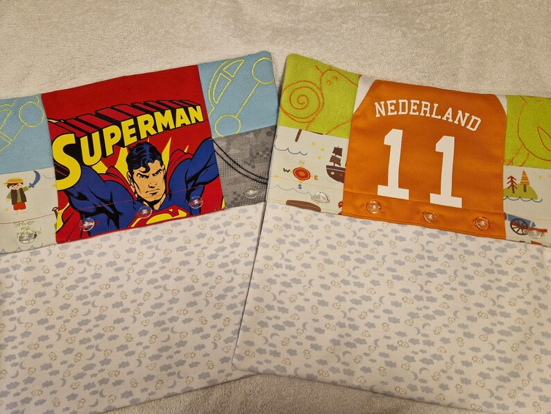 May include: Two rectangular fabric pillowcases featuring patchwork designs. The left case displays Superman with yellow text, while the right case features an orange panel with the text NEDERLAND 11. Both have white sections with small cloud and moon patterns.