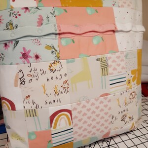 May include: A square patchwork cushion made from various fabric scraps featuring patterns of rainbows, pears, hedgehogs, snails, and giraffes. The surface includes horizontal strips of light blue and pink fabric with integrated snap buttons.