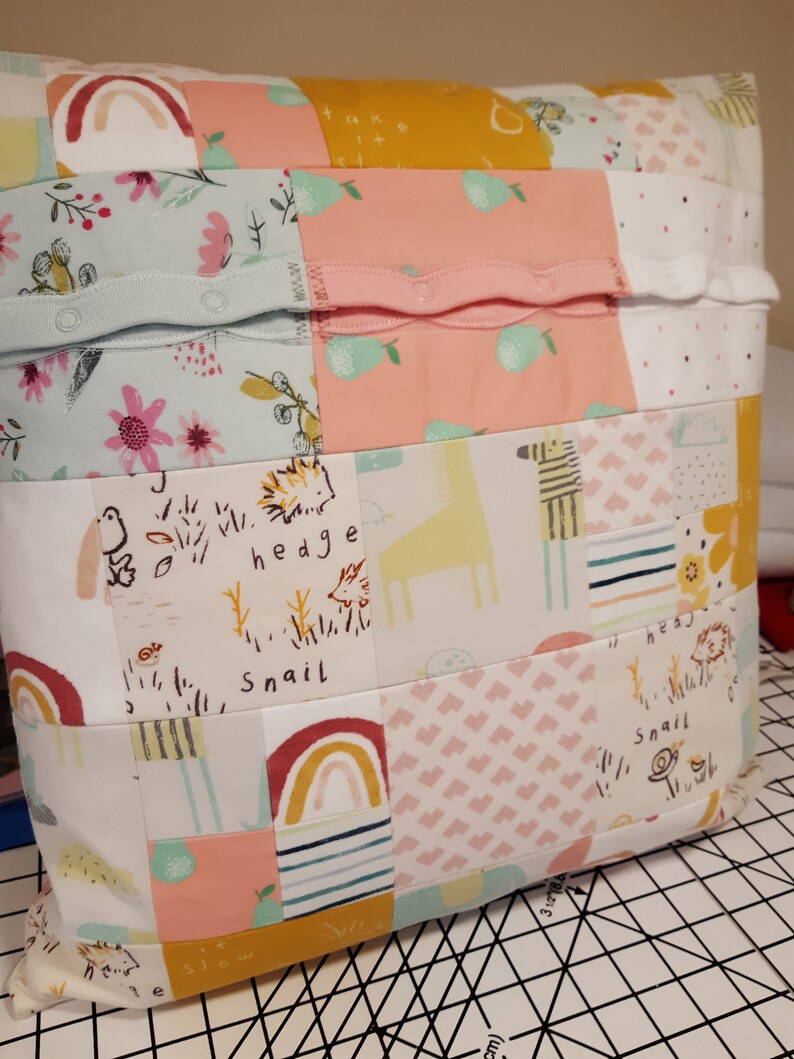 May include: A square patchwork cushion made from various fabric scraps featuring patterns of rainbows, pears, hedgehogs, snails, and giraffes. The surface includes horizontal strips of light blue and pink fabric with integrated snap buttons.
