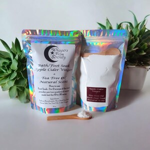 May include: Two holographic pouches of bath and foot soak powder with a wooden scoop. The pouches are labeled "Whispers From Serenity" and list ingredients and instructions. The product is Apple Cider Vinegar and Tea Tree Oil scented.