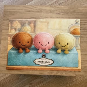 Peut inclure: Illustration fantaisiste de trois personnages ronds et souriants en peluche marron, rose et jaune, avec de petites jambes, assis sur une boîte bleue. La boîte porte l'inscription "Patisserie". L'arrière-plan montre une scène de boulangerie.
