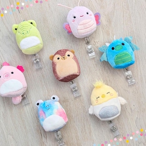 May include: A collection of plush badge reels in various animal shapes, including a frog, butterfly, monkey, dragon, pig, and chick. Each reel is a different color and has a retractable cord and a clear plastic clip.