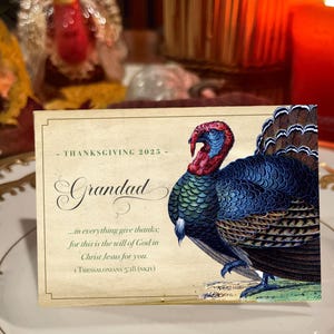 Op de afbeelding: Thanksgiving 2025 wenskaart met een gedetailleerde illustratie van een kalkoen met blauwe, rode en bruine veren. De kaart leest "Grandad" en bevat een bijbelvers. De kaart is rechthoekig.