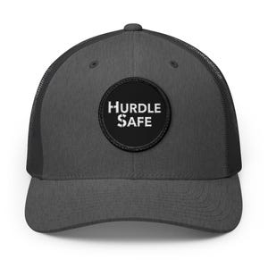 May include: A charcoal gray trucker hat featuring a black circular patch on the front panel. The patch displays the white text HURDLE SAFE in a distressed, bold font. The hat has a structured front, a curved brim, and a mesh back panel.