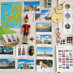 Könnte beinhalten: Eine Sammlung von Lernspielkarten mit Bildern europäischer Sehenswürdigkeiten, Flaggen und kultureller Symbole. Die Karten zeigen Bilder vom Eiffelturm, Big Ben, dem Kolosseum und der Basilius-Kathedrale. Es gibt auch Karten, die traditionelle Kleidung aus verschiedenen europäischen Ländern wie der Ukraine und Portugal zeigen. Die Karten sind auf einer weißen Oberfläche mit einer Holzpuppe, einem Holz-Nussknacker und einem grünen Umschlag angeordnet.