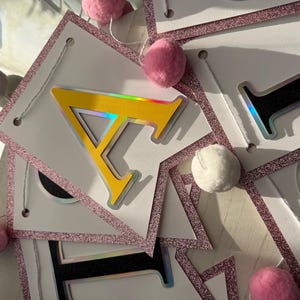 May include: A banner with white pennants, each outlined with pink glitter and strung together with white string. The letter "A" is yellow with a silver outline. Pink and white pom-poms are interspersed along the string.