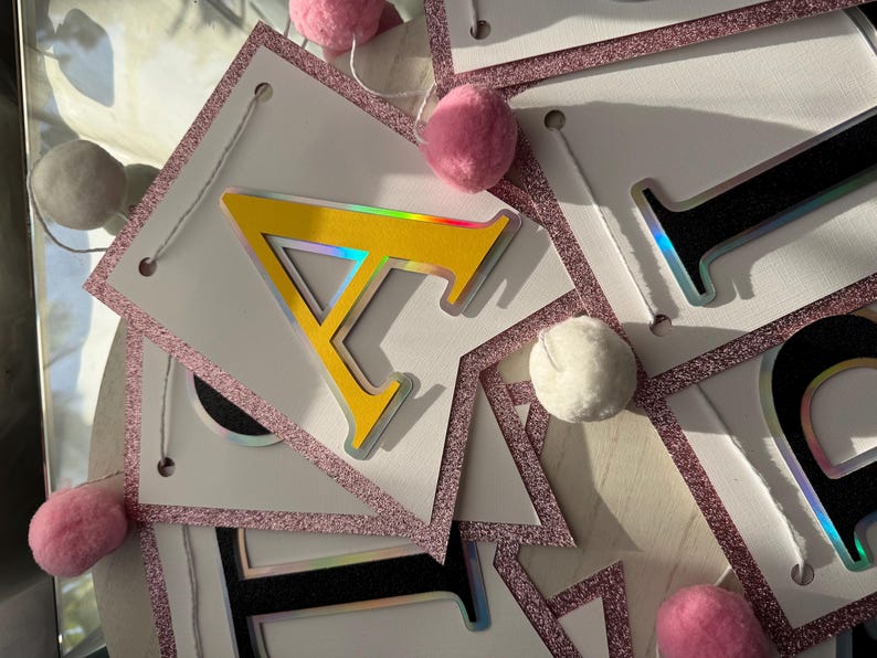 May include: A banner with white pennants, each outlined with pink glitter and strung together with white string. The letter "A" is yellow with a silver outline. Pink and white pom-poms are interspersed along the string.