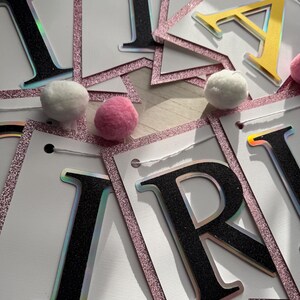 May include: A banner with individual letter flags spelling out a word. The letters are black with a holographic edge, and the flags have a pink glitter border. White and pink pom-poms are scattered around the banner.