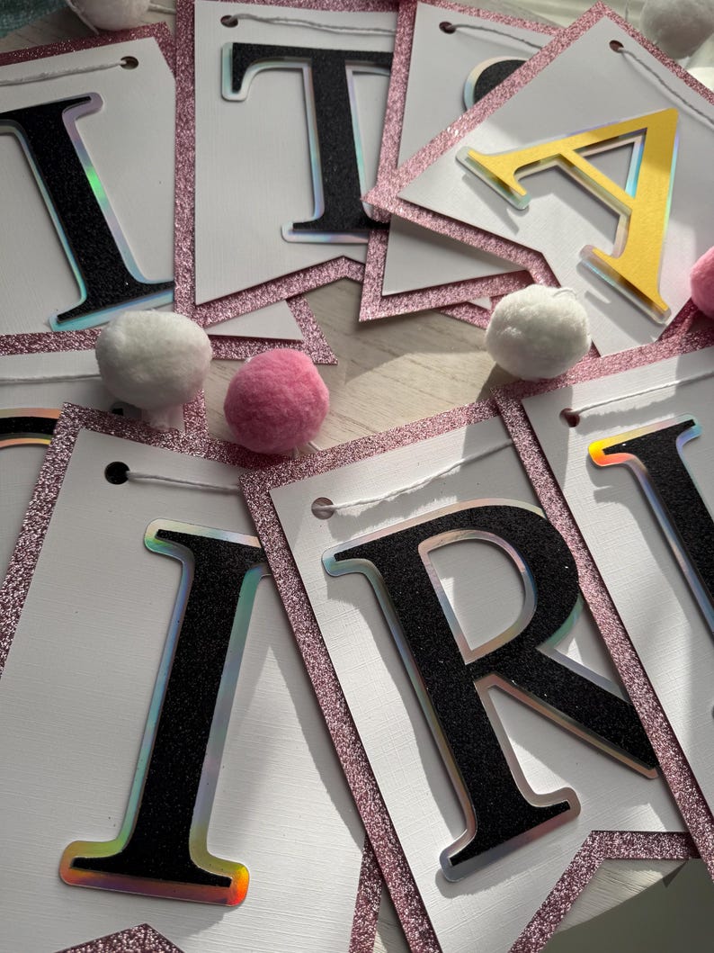 May include: A banner with individual letter flags spelling out a word. The letters are black with a holographic edge, and the flags have a pink glitter border. White and pink pom-poms are scattered around the banner.