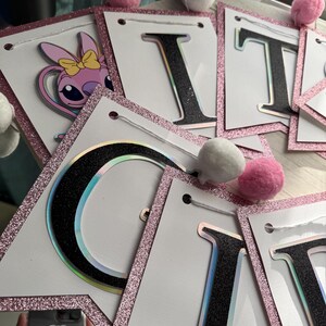 May include: A banner spelling out "IT'S A GIRL" with white pennants, black glitter letters, and pink glitter borders. The banner includes a cartoon character and is decorated with pink and white pom-poms.