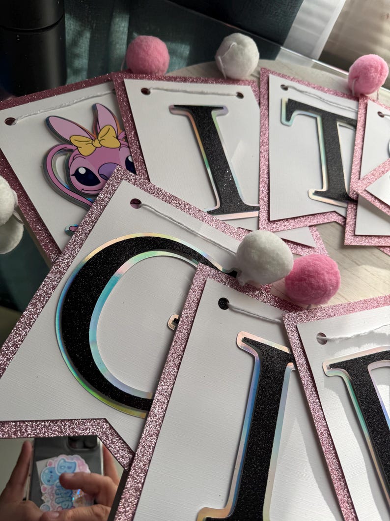 May include: A banner spelling out "IT'S A GIRL" with white pennants, black glitter letters, and pink glitter borders. The banner includes a cartoon character and is decorated with pink and white pom-poms.