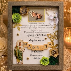 May include: A rectangular gray wooden shadow box frame containing a photograph of a baby, a small rosary, paper flowers in green and gold, a gold butterfly cutout, a religious figure, and wooden letter tiles spelling MADRINA in Spanish.