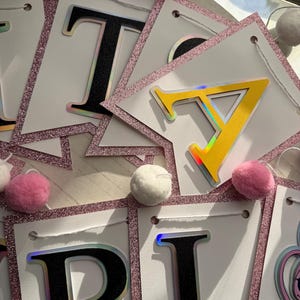 May include: A banner spelling out "TARA" with white pennants, black letters with iridescent outlines, and a pink glitter border. Pink and white pom-poms are scattered around the banner. The banner is strung with white string.