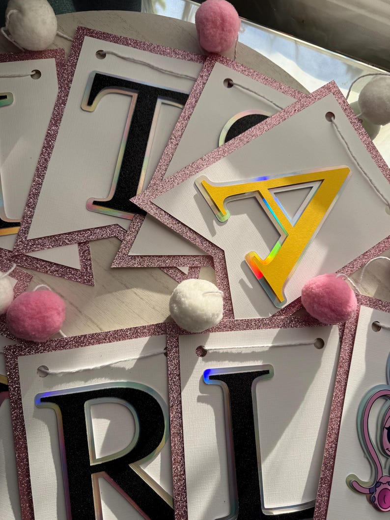 May include: A banner spelling out "TARA" with white pennants, black letters with iridescent outlines, and a pink glitter border. Pink and white pom-poms are scattered around the banner. The banner is strung with white string.