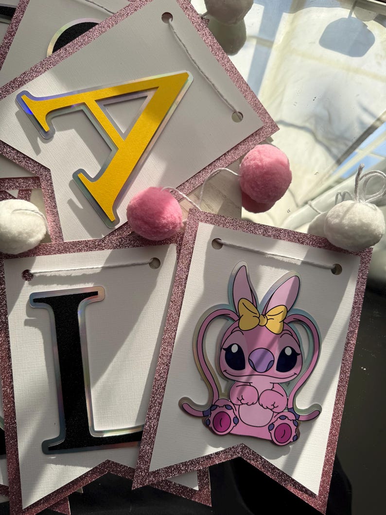 May include: A banner with white pennants, each outlined with pink glitter. The banner spells out "I LOVE" with yellow and black letters. A pink cartoon character with a yellow bow is featured on one pennant, and pink and white pom-poms are attached.