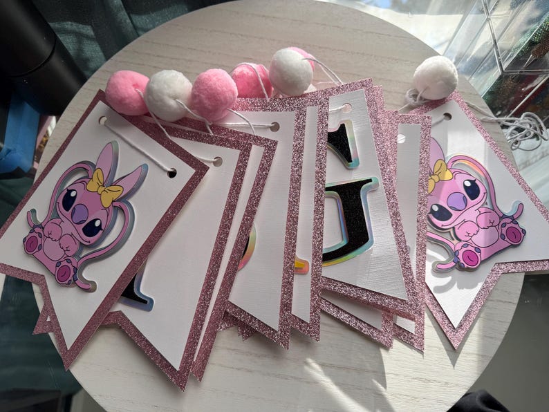 May include: A pink and white banner with glitter trim and pom-poms. Each pennant features a cartoon character with a yellow bow. The banner spells out "1st" with a cartoon character on each side.