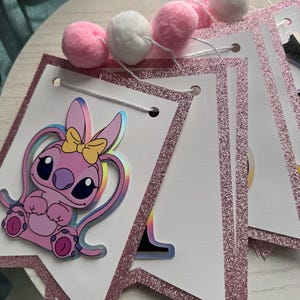 May include: A pink and white banner with a glittery pink border and a cartoon character illustration. The banner is strung with white string and adorned with pink and white pom-poms. The cartoon character has a yellow bow.