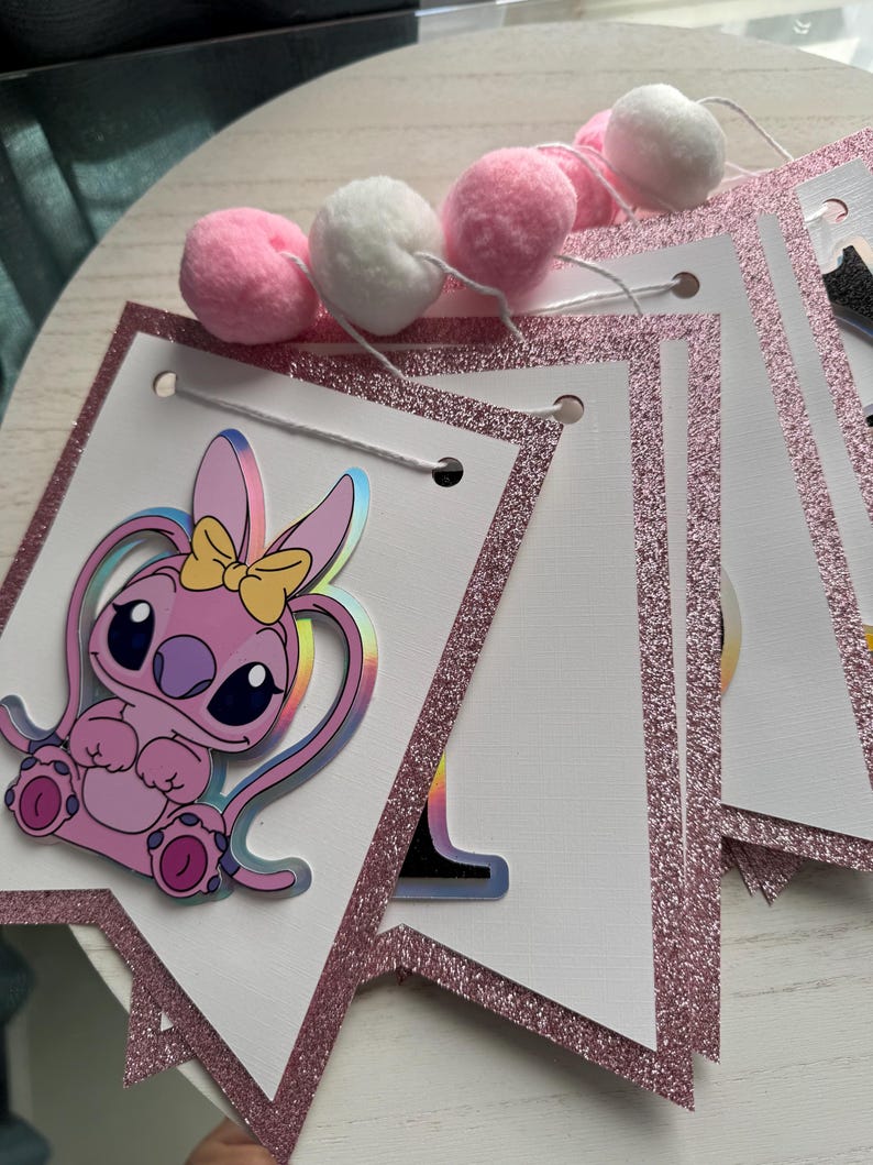May include: A pink and white banner with a glittery pink border and a cartoon character illustration. The banner is strung with white string and adorned with pink and white pom-poms. The cartoon character has a yellow bow.