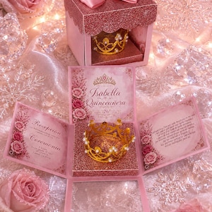 May include: A pink glittery gift box with a satin bow and a gold crown inside. The box unfolds to reveal printed text including Isabella Quinceañera, August 26, 2023, and event details for a Recepción and Ceremonia, decorated with pink rose illustrations.