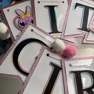 May include: A banner with white pennants, each outlined with pink glitter. The letters are black with a holographic effect. One pennant features a cartoon character. Pink and white pom-poms are scattered around the banner.