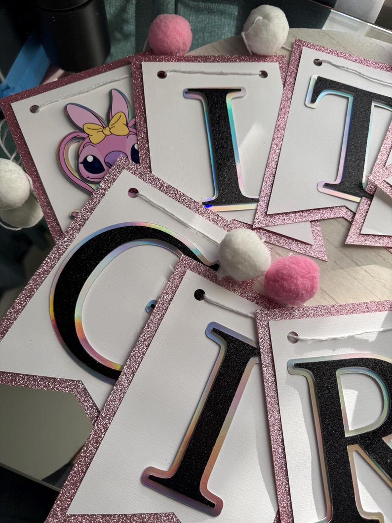 May include: A banner with white pennants, each outlined with pink glitter. The letters are black with a holographic effect. One pennant features a cartoon character. Pink and white pom-poms are scattered around the banner.