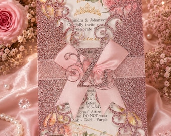 May include: A rectangular invitation card featuring a rose gold glitter laser-cut cover with intricate scrollwork. A light pink satin ribbon is tied across the center, accented by a glittery number 21. The card displays printed text and a gold crown.