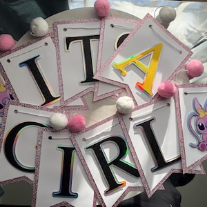 May include: A banner that spells out "IT'S A GIRL" with individual pennants. Each white pennant has a pink glitter border and is strung together with string. The letters are black with a holographic effect. Pink and white pom-poms are attached.
