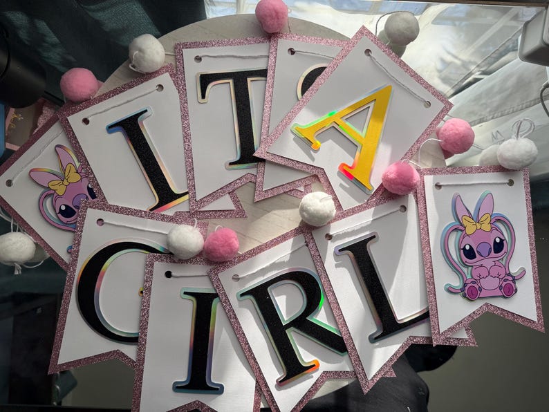 May include: A banner that spells out "IT'S A GIRL" with individual pennants. Each white pennant has a pink glitter border and is strung together with string. The letters are black with a holographic effect. Pink and white pom-poms are attached.