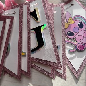 May include: A pink and white banner with glittery pink borders and a string of pink and white pom-poms. The banner features letters with holographic accents and a cartoon character.