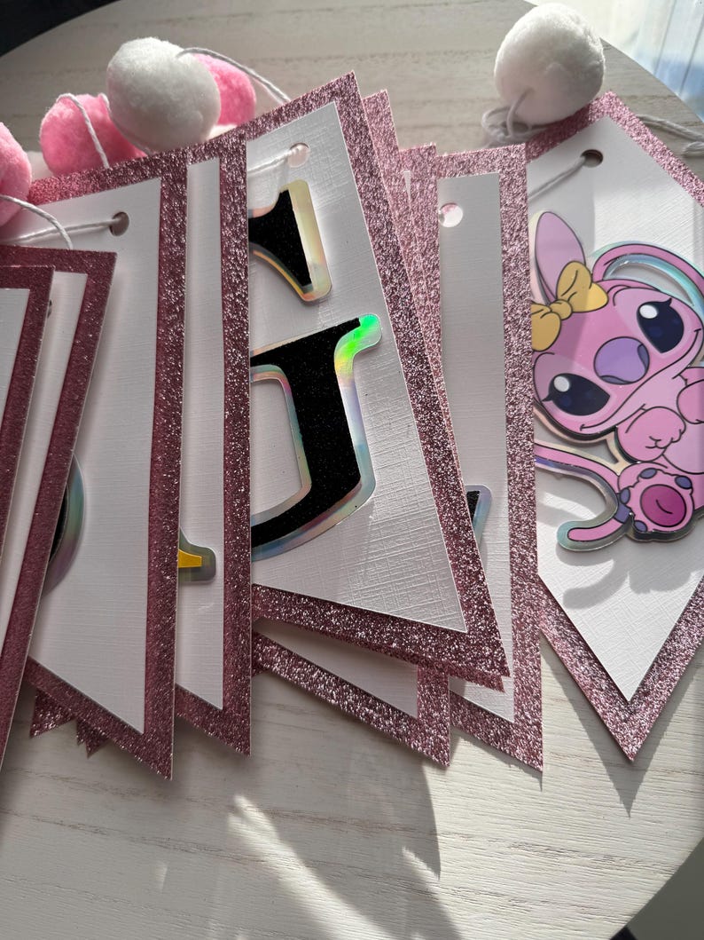 May include: A pink and white banner with glittery pink borders and a string of pink and white pom-poms. The banner features letters with holographic accents and a cartoon character.