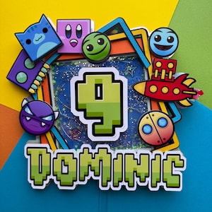 May include: A colorful cake topper with the name "DOMINIC" in pixelated green letters. The number "9" is in the center, surrounded by various cartoon characters and a red rocket ship. The background is a mix of blue, green, and yellow.