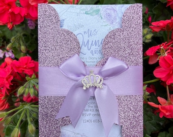 May include: A lavender glitter invitation with a light purple ribbon and a silver crown charm. The invitation has the words "Mis Quince años" printed on the front. The background features a floral design.