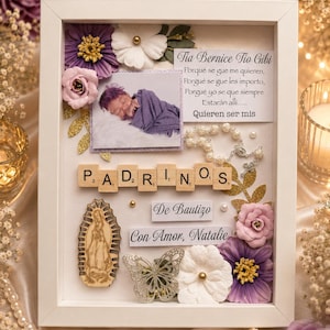 May include: A white shadow box frame containing a photograph of a swaddled infant, wooden letter tiles spelling PADRINOS, a small rosary, a wooden Virgin Mary cutout, a butterfly cutout, and paper flowers with the text Tia Bernice Tio Gibi.