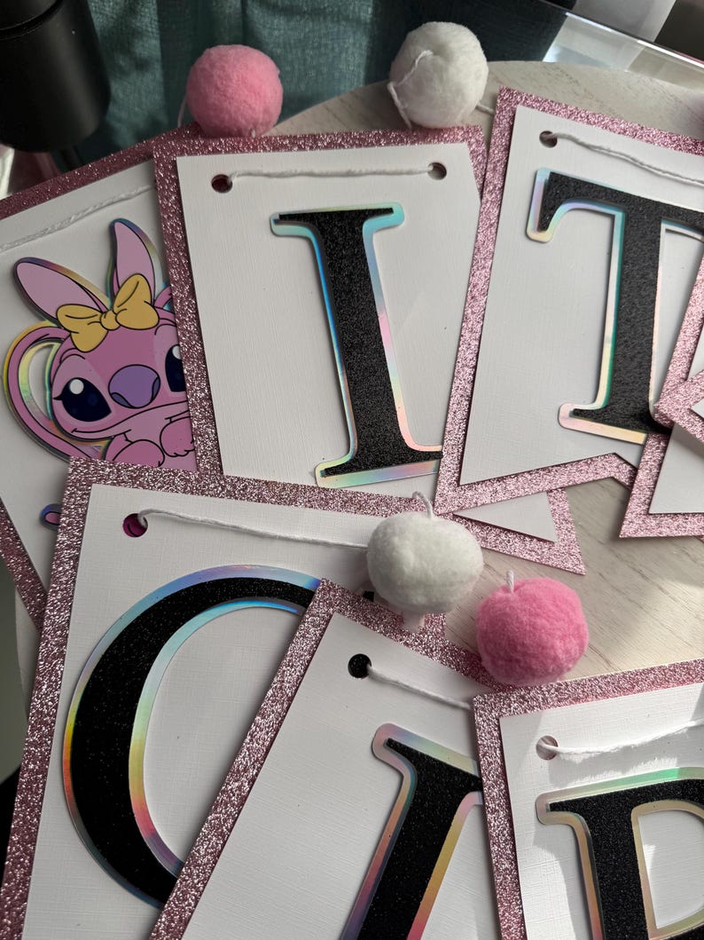 May include: A banner with white pennants, outlined with pink glitter, and black letters with a holographic effect. The banner is decorated with pink and white pom-poms. A pink cartoon character with a yellow bow is visible.