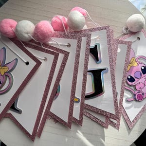 May include: A pink and white banner with glitter trim and pom-poms. The banner features a cartoon character and the letter "J". The pom-poms are pink and white. The banner is likely for a birthday or party.
