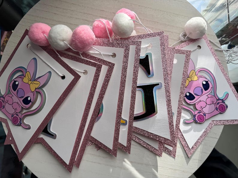 May include: A pink and white banner with glitter trim and pom-poms. The banner features a cartoon character and the letter "J". The pom-poms are pink and white. The banner is likely for a birthday or party.