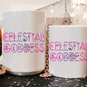 May include: Two white ceramic mugs with the words "CELESTIAL GODDESS" in a gradient pink and purple font. The design includes celestial symbols like moons, suns, and stars. A gold chain hangs between the mugs.