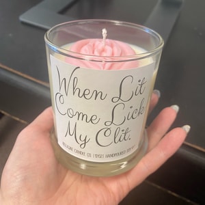 May include: A white candle in a glass jar with a pink wax flower on top. The jar has a white label with the text "When Lit Come Lick My Clit." and the text "BELLALJAE CANDLE CO | 12 OZ | HANDPOURED SOY."