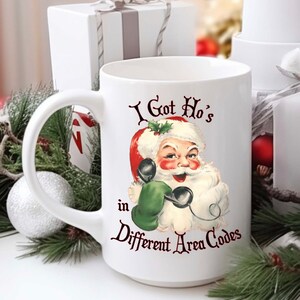 May include: White ceramic mug with a red and white illustration of Santa Claus on the phone. The text on the mug reads "I Got Ho's in Different Area Codes".