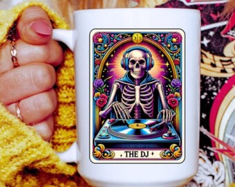 May include: White ceramic mug featuring a colorful illustration of a skeleton DJ wearing headphones and operating a turntable. The design includes the text "THE DJ" and floral accents. The mug is held by a person wearing a yellow sweater.
