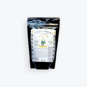 Op de afbeelding: Een zwarte hersluitbare zak met een wit etiket waarop "SCOTT FARMS" en "CORN CHOWDER SOUP MIX" staat. Het etiket bevat ook een cartoonafbeelding van een maïskolf met een zwaard. De zak bevat 9 oz (255 gram) maïs soepmix.
