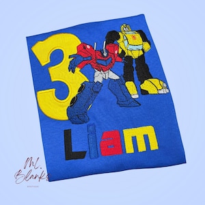 Peut inclure: Un t-shirt bleu avec une broderie représentant un grand chiffre 3 jaune, un robot rouge et bleu, et un robot jaune et noir. Sous les robots, le prénom Liam est brodé en lettres capitales noires, bleues, rouges et jaunes.